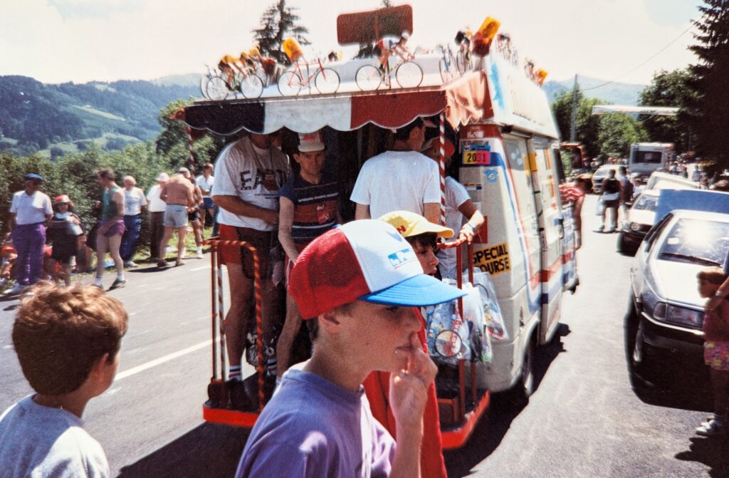 A colorful publicity van drives on a road in the Alps during the 1990 Tour de France. The van is adorned with a red, white, and blue canopy and miniature bicycles perched on top. People in casual summer attire, some with hats, stand on an open platform off the back of the van. Some young boys are in the foreground. The background features tree-covered hills beneath a partly cloudy sky, giving the scene a festive, small-town vibe.