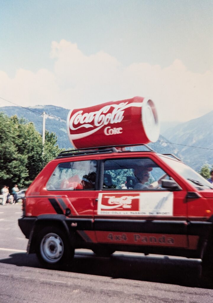 A red 4x4 Fiat Panda with an oversized Coke can mounted on its roof rack drives by. In the background are trees and distant Alpine mountains.