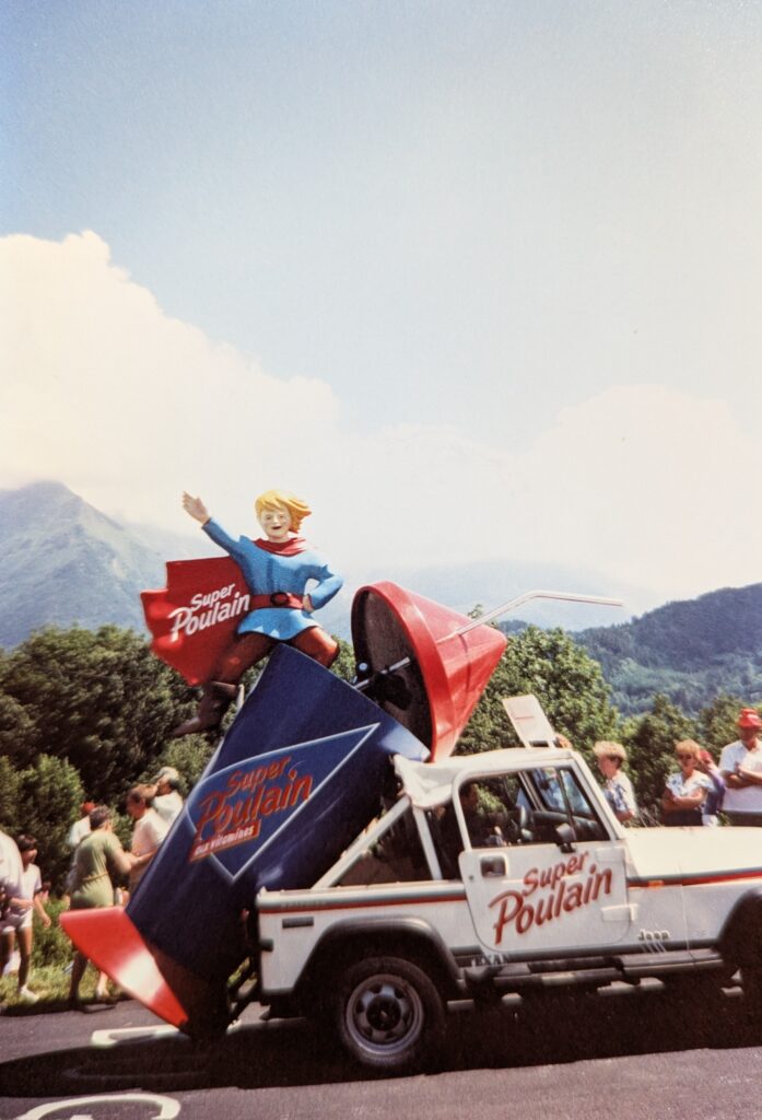 A white Jeep Wrangler with a thick red pinstripe drives uphill in the Alps. The passenger door has "Super Poulain" painted in bright red letters. In back, the rear roof of the Jeep has been removed so that an oversized - maybe 10 feet - drinking cup can be mounted. The cup - blue with "Super Poulain" written on it, hangs off the back of the Jeep, the red base of the cup nearly touching the ground. The oversized conical red lid and straw are tilted off the cup and rest on the roof. A cartoon character - a young blond boy with red tights, blue tunic, red belt, and red cape emerges from the cup. On his cape, "Super Poulain" is written in white.