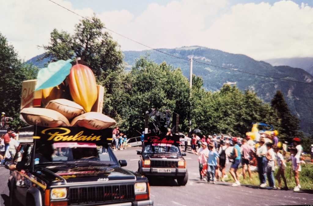 Photo looking down towards a hairpin curve on a mountain road in the French Alps. It is summer. People are lining both sides of the road as two Jeep Cherokees drive up through the hairpin. Each Jeep has an oversized marketing display on the roof. The first has a sign "Poulain" above the windshield, with oversized sacks of cacao beans, an even larger cacao bean and leaf, and retail packaging. The other is also "Poulain" but the objects on top are black and difficult to discern.