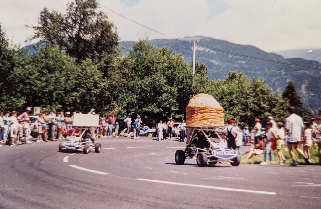 A group of spectators lines a curved mountain road in the Alps during the Tour de France, watching a pair of whimsical go-karts drive up a hairpin turn. One go-kart features a large prop shaped like a loaf of bread, while another sports a rectangular box. Both vehicles are labeled “PATIGEL.” The scene is festive, with people casually gathered on the roadside and lush hills rising in the background.