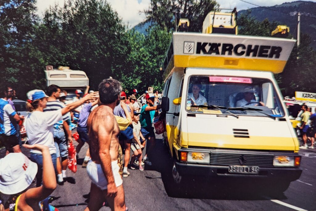 A promotional RV from the company Karcher drives up the hairpin turn. The RV is yellow and white with a matching yellow and white display on the roof with the name and logo Karcher prominently displayed. On top appears to be a yellow and white generator and scale model construction equipment in yellow. People line the side of the road and reach out to the Karcher RV.