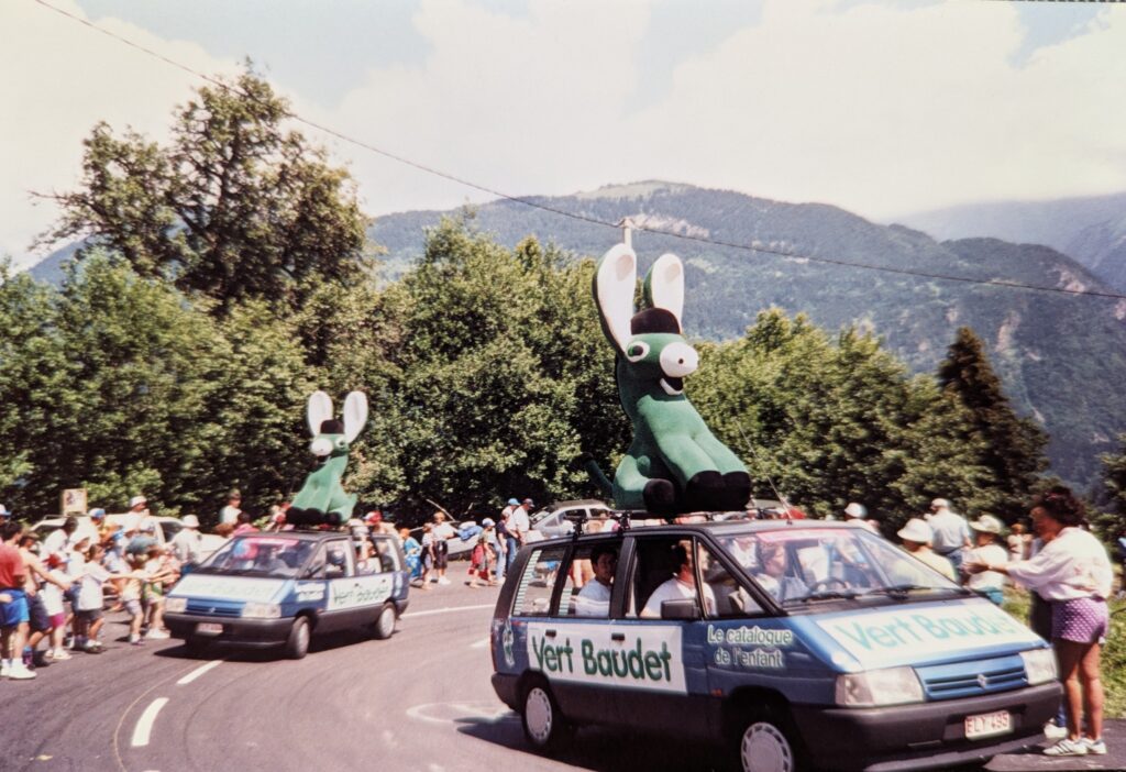 Two blue Renault minivans drive up through a hairpin turn in the Alps. The roads are lined with people waiting for the Tour de France. The vans have white placards with the words "Vert Baudet" written in large green letters along their sides and hood. On top of the vans are giant green oversized donkey-like figures. They are seated on their haunches, their ears are up, they are leaning back, and the expression on their faces suggest that they are excited to be moving fast.