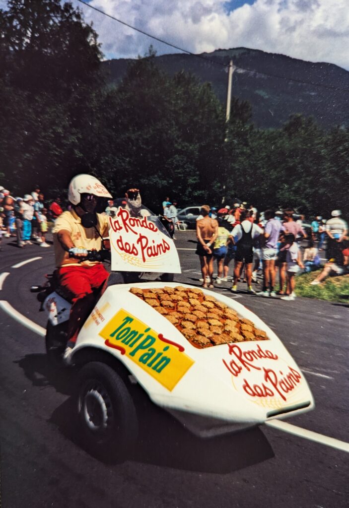 A lively outdoor scene on a mountain road in the Alps shows a person in a yellow shirt and red pants rides a tricycle - a motorcycle that has been modified with two front wheels to carry a white car-like nose. The nose is decorated with French text promoting bread and "La Ronde des Pains." and on top has what appear to be biscuits or some kind of bread. Behind the motorcycle, crowds of spectators line the road. Lush green trees and dramatic mountains rise in the background under a partly cloudy sky.