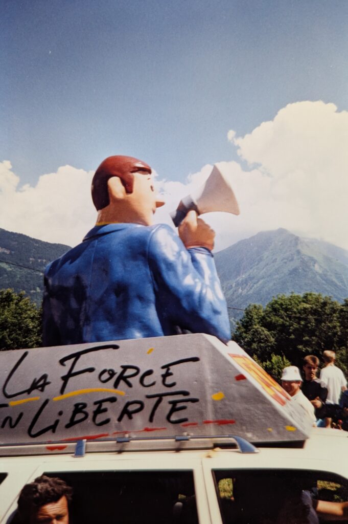 A colorful parade float features a large cartoonish figure of a man in a blue jacket holding a megaphone, mounted on top of a vehicle. The side of the float displays the French words "La Force en Liberté" in bold, playful lettering. The scene is set outdoors with green trees and mountains in the background under a bright, partly cloudy sky. Several people are gathered around the float, enjoying the event