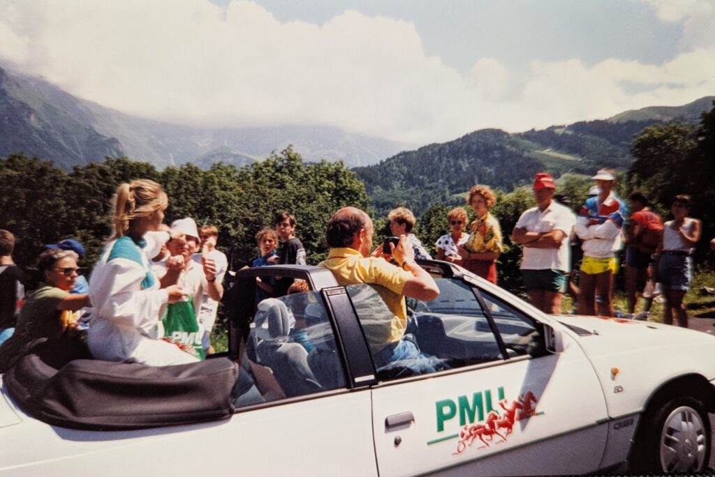A slightly elevated outdoor shot captures a white convertible car driving uphill with a crowd of people gathered around it under a partly cloudy sky. The car is angled from the front passenger side, with its top down. In the front passenger seat, a person with short brown hair, wearing a yellow collared shirt, is lifting himself above the windshield and taking a picture with a camera held to their eye, facing towards the right side of the frame. In the back seat on the left, a blonde-haired woman in a white and teal top is seated on the folded convertible top, her head above the rollbar The car has a green "PMI" logo with two red stylized horse figures on its front passenger door. Numerous spectators are visible around the car. On the left side of the frame, several people are standing near the car. On the right side of the car, a larger group of people of varying ages and attire are standing and looking towards the general direction of the car or further into the background. The background features lush green trees and a mountainous landscape under a bright sky with scattered clouds. The mountains are hazy in the distance. The overall impression is of a public event, possibly a race or parade, where spectators are eagerly watching.