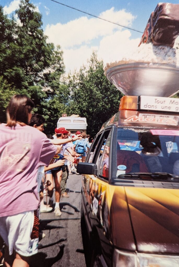 A bright, sunny, eye-level outdoor shot shows a car, angled from the front passenger side, in the foreground of a gathering of people. The car, which is brown fading into dark yellow, has an open window on the passenger side. On its roof, there is a large, silvery bowl or dish, filled with a white, fluffy substance, possibly ice, and what appear to be a large chocolate bar sticking out of it. Underneath the bowl is a light brown rectangular box with a yellow flashing light. On the side of the box, text in a foreign language is visible, with a few words visible that read "Tout Mo" and "dans une g." Below is the "Mars" logo.. To the left, a person with medium-length brown hair, wearing a purple t-shirt and white shorts, has their back to the viewer and is extending their right arm towards the car. In the background, many people are walking around and standing. One person in a red cap is prominent, while others are less distinct. Beyond them, a few white trailers or trucks are visible. The scene is framed by tall green trees on both sides, and a bright blue sky with scattered white clouds is overhead. The overall impression is one of a lively, outdoor event.