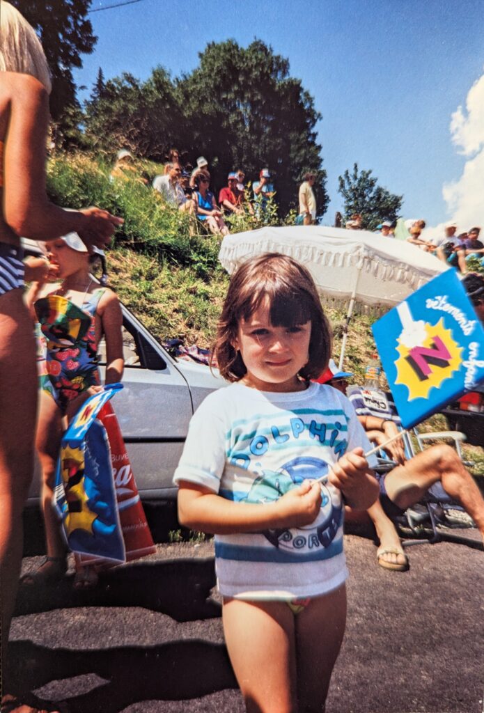 A young girl stands in the foreground holding a small, vibrant flag from the cycling team Z. The child wears a white T-shirt with blue accents and the word "DOLPHIN" across the front. Behind them, people are relaxing on a grassy hill, some shaded under a white umbrella, with a parked car nearby. The setting is sunny and outdoors, on an Alpine roadside at the Tour de France