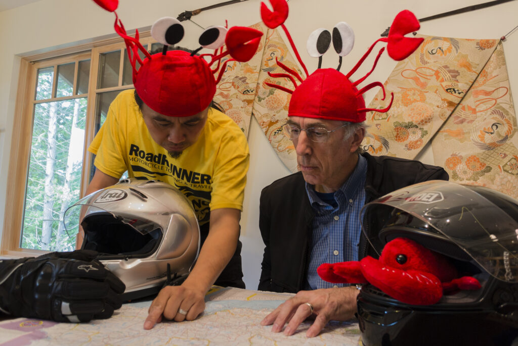Two men study a map that's spread out on the table in front of them. On the table are two motorcycle helmets and a pair of motorcycle gloves. A red lobster plushy sits inside one of the motorcycle helmets. The two men are wearing silly red lobster hats with decorative felt claws, googly eyes, and tentacles.