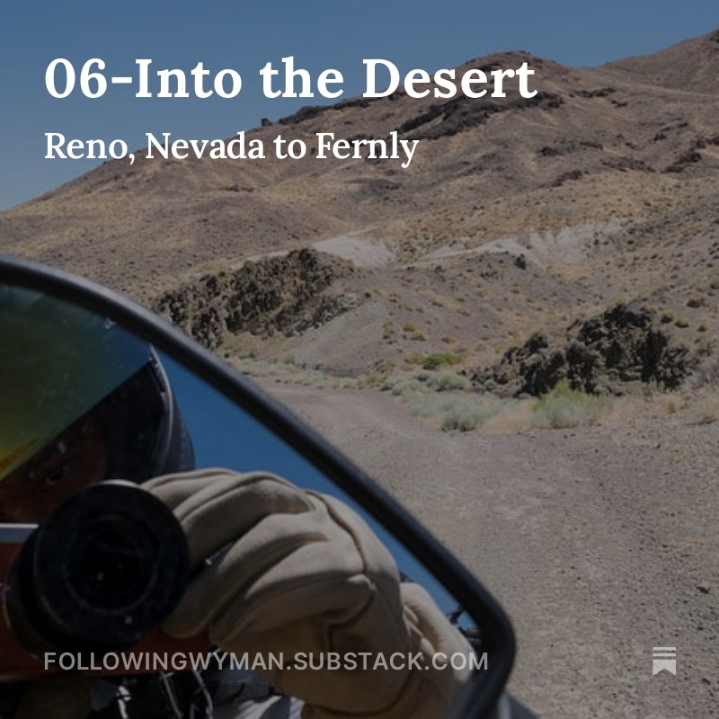 Photo from the seat of a motorcycle, including the land ahead of the motorcycle, a piece of the windshield on the right edge of the photo, and the author reflected in the left rearview mirror in the lower left corner of the photo. The land ahead of the motorcycle is a tall, naked mountain in the desert with multiple, weather-worn peaks leading up to a naked top. Small clumps of grass grow in patches and exposed, dark rock appear on the hill in places. The dirt road that the motorcycle on bend to the left. The author reflected in the mirror of the motorcycle is wearing elkskin leather gloves and a black helmet with a checkerboard strip pattern along the top and a gold reflective visor that has been flipped up a little for ventilation. The reflection in the visor is more scrub brush growing in the sandy soil and the blue sky, both tinted gold. The author is holding a small orange camera with a black lens that is being used to take the photo. Headline at top 06-Into the Desert Subhead Reno, Nevada to Fernly Footer FollowingWyman.Substack.com