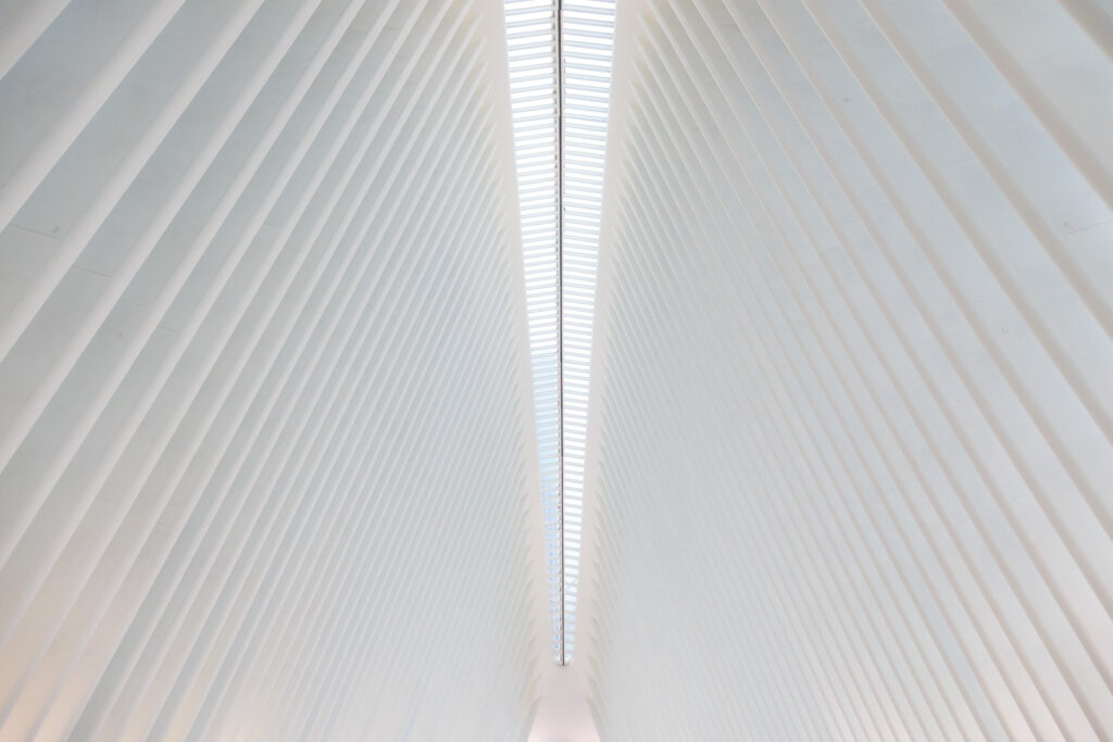 A photo looking up at the shimmering white ceiling of the Oculus Transportation Hub in New York, designed by Spanish architect Santiago Calatrava. The ceiling is comprised of closely spaced white ribs rising up towards a clear glass skylight that arcs over the center of the frame towards a terminus point at the far end of the hall. It kind of feels like being in a giant whale; the skylight is the spine and the white ribs define the space.