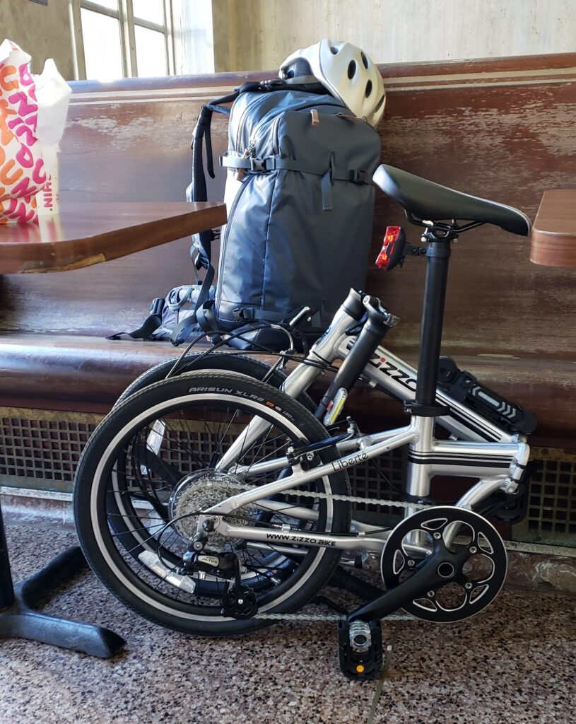 Photo of a folding bicycle folded up and on the floor of a train station. Behind the bike is an old wooden bench where there's a blue backpack with a bicycle helmet sitting on it.
