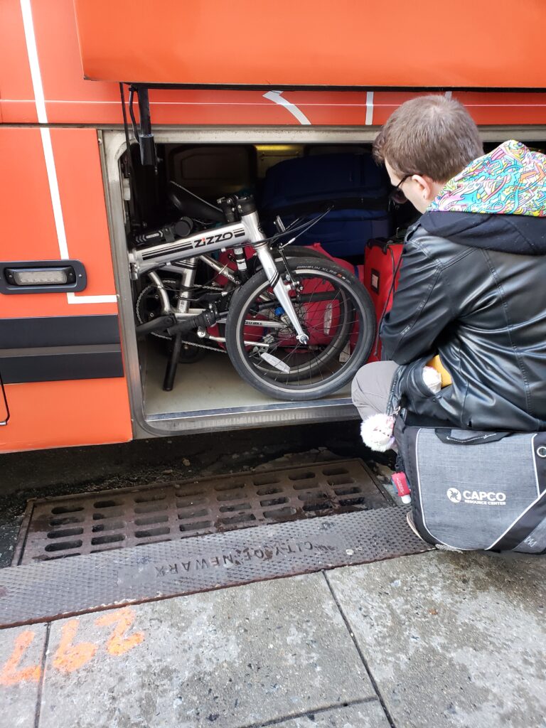 Photo of a folded up folding bicycle in the below storage area of a bus. The bike is surrounded by luggage. A man is placing his luggage in the storage area.