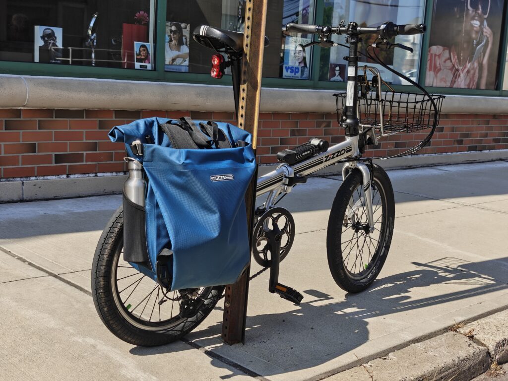 Photo of a folding bike locked to the pole of a street sign. Attached to the rear rack is a blue bag.