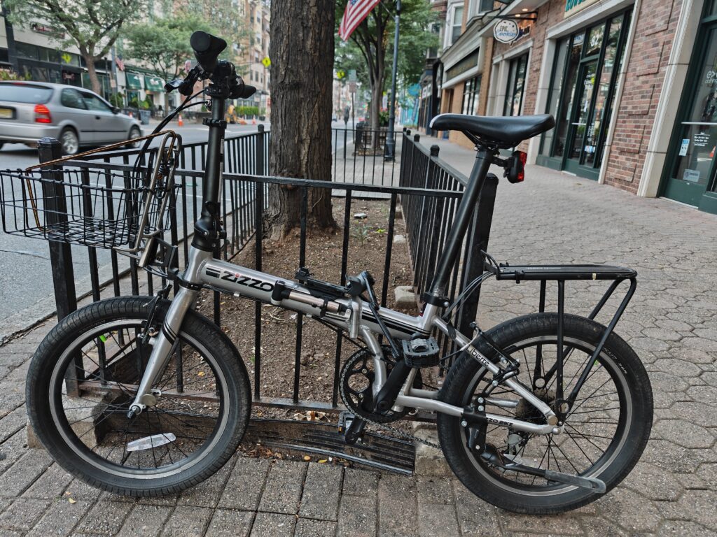 Photo of a silver folding bicycle locked to a wrought iron railing on the sidewalk of a downtown area