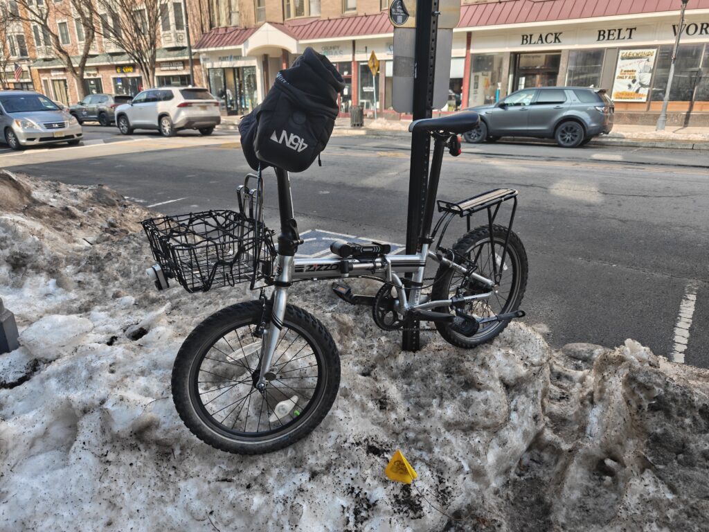 Photo of a folding bicycle locked to a street sign in winter. The bike is sitting upon an icy snowbank. There are warming pogies on the handlebars