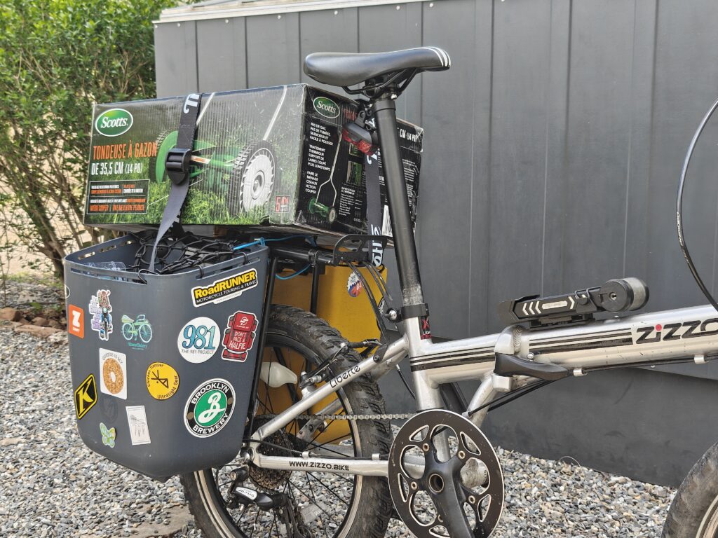 Photo of the rear half of a folding bicycle with plastic bucket panniers. On top of the rear rack is a large box held down with a wide strap. The box has a photo of its contents - a manual push reel mower.