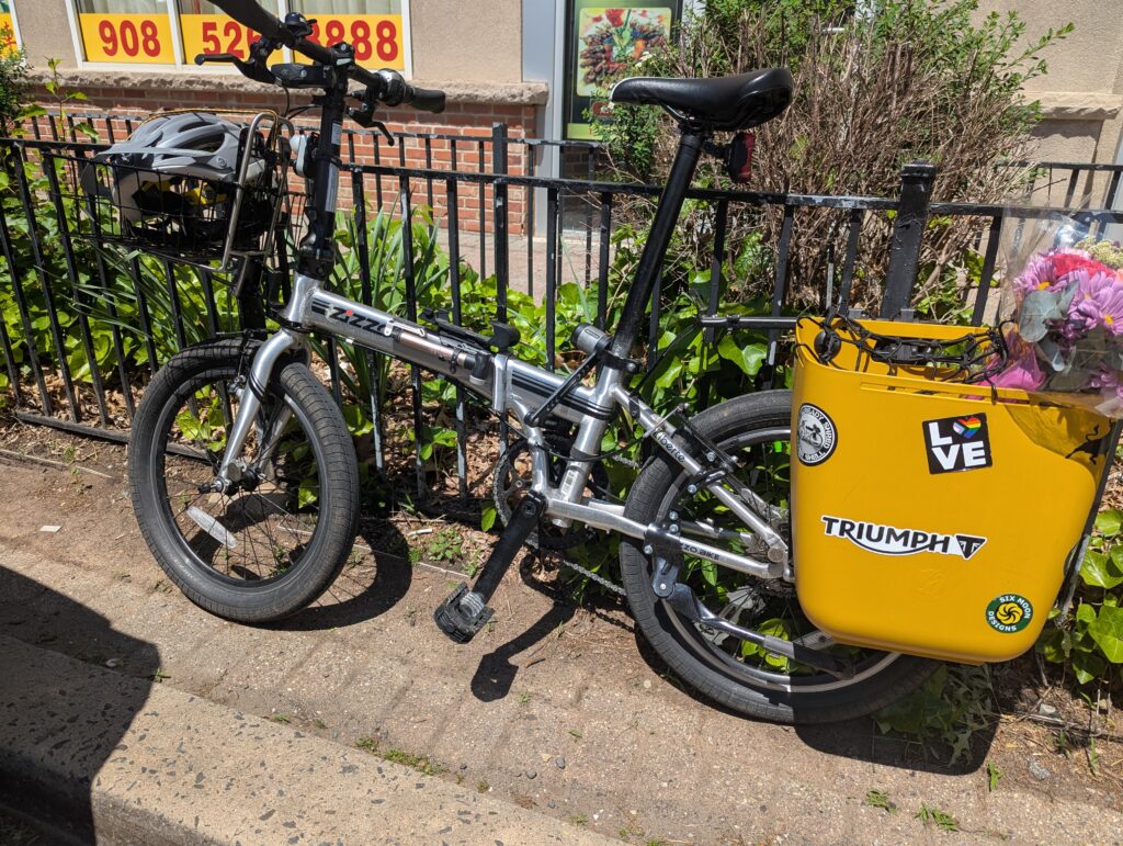 Photo of a silver folding bike locked to a wrought iron rail. In the front basket is a helmet. In the rear plastic bucket pannier is a bouquet of flowers.