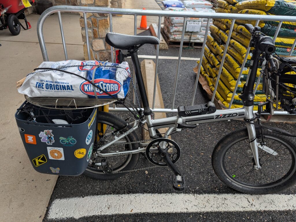 Photo of a folding bicycle with plastic panniers parked outside a home improvement store. On the rear rack of the bicycle is a round grill and a bag or charcoal briquets. They are tied down with straps.