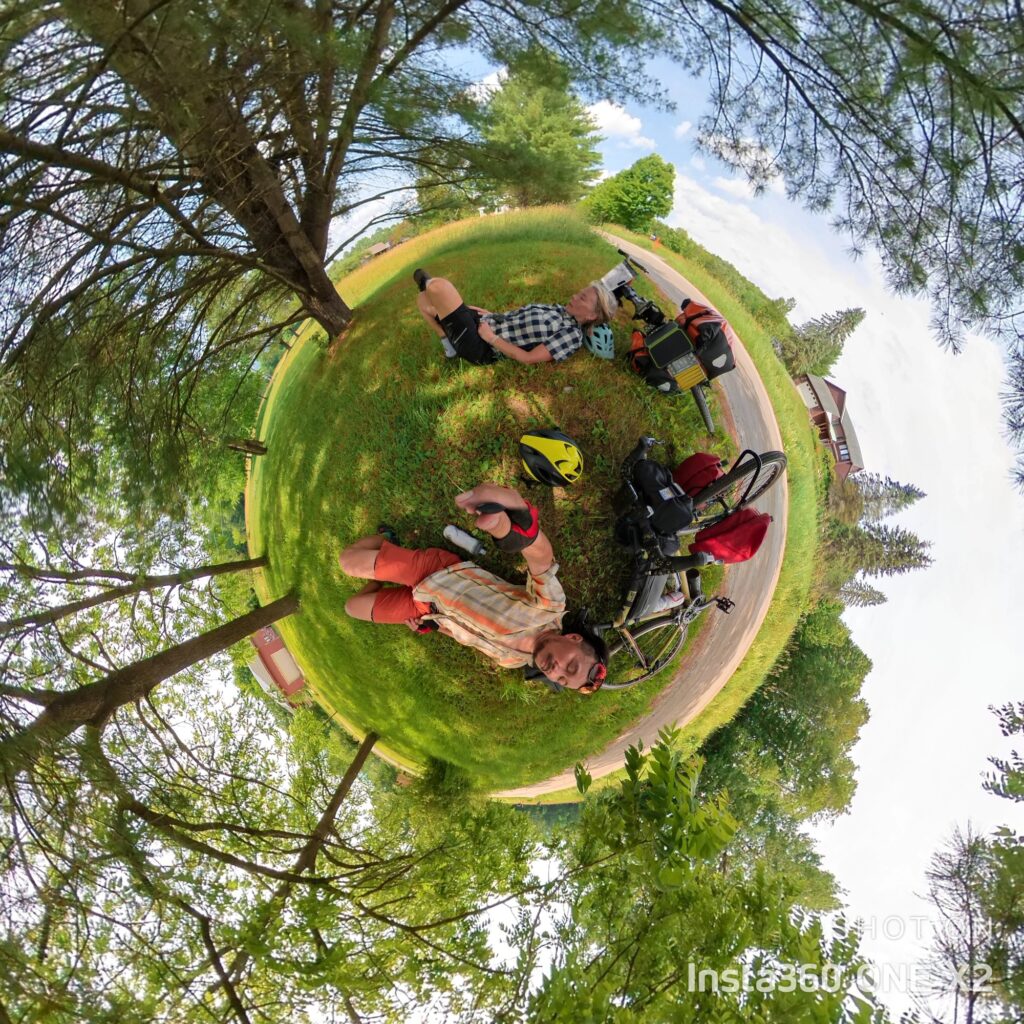 Photo of two bicycle riders taking a break under the shade of a tree in Vermont The bicycles with their bags are laying on the grass as they lay beside them, fatigued