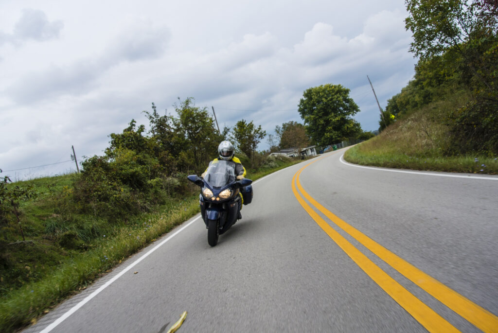 Photo of a man on a blue motorcycle in the middle of a curve on a country road. The motorcycle is leaning dramatically in the turn. There are trees and shrubs on the side of the road. The sky is cloudy.