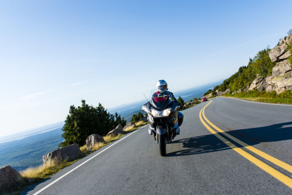 A man rides a motorcycle up a winding road on a mountain. In the distance and below the mountain are forests, and to the right of the road is the stony mountain. The rider is leaning the bike into the turn. The sky is blue and hazy