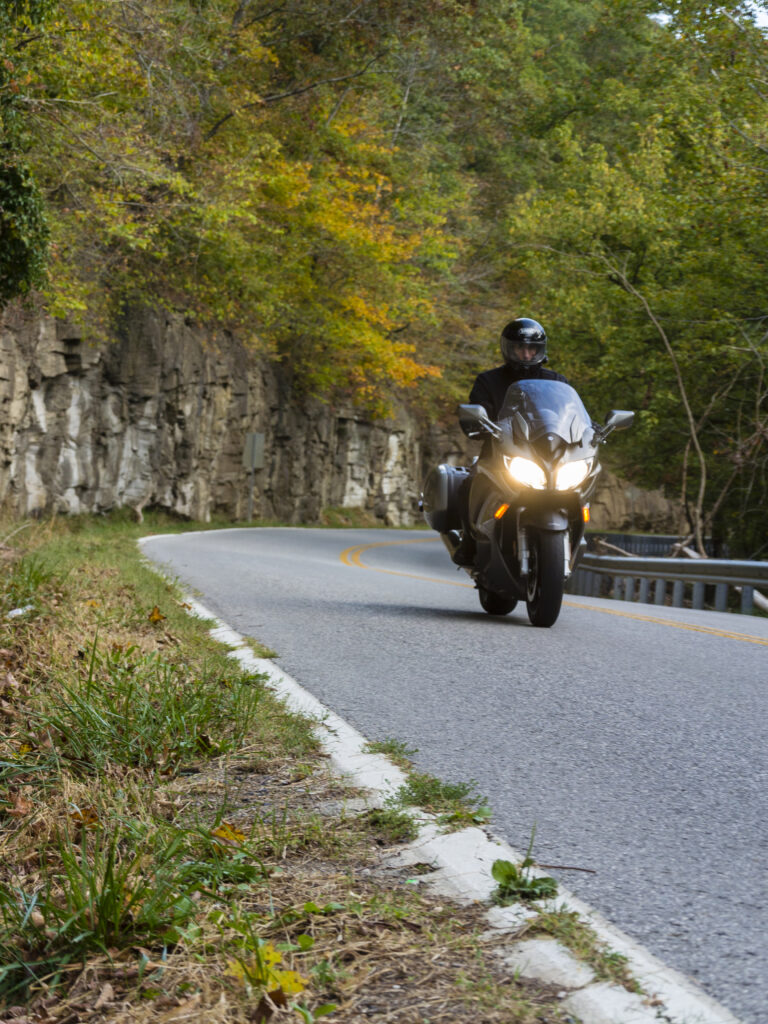 A man rides a motorcycle on a curvy country road. Along the side of the 2-lane narrow road is a rock wall rising ~20 feet from the ground. Beyond are lush green trees with some of the leaves turning yellow