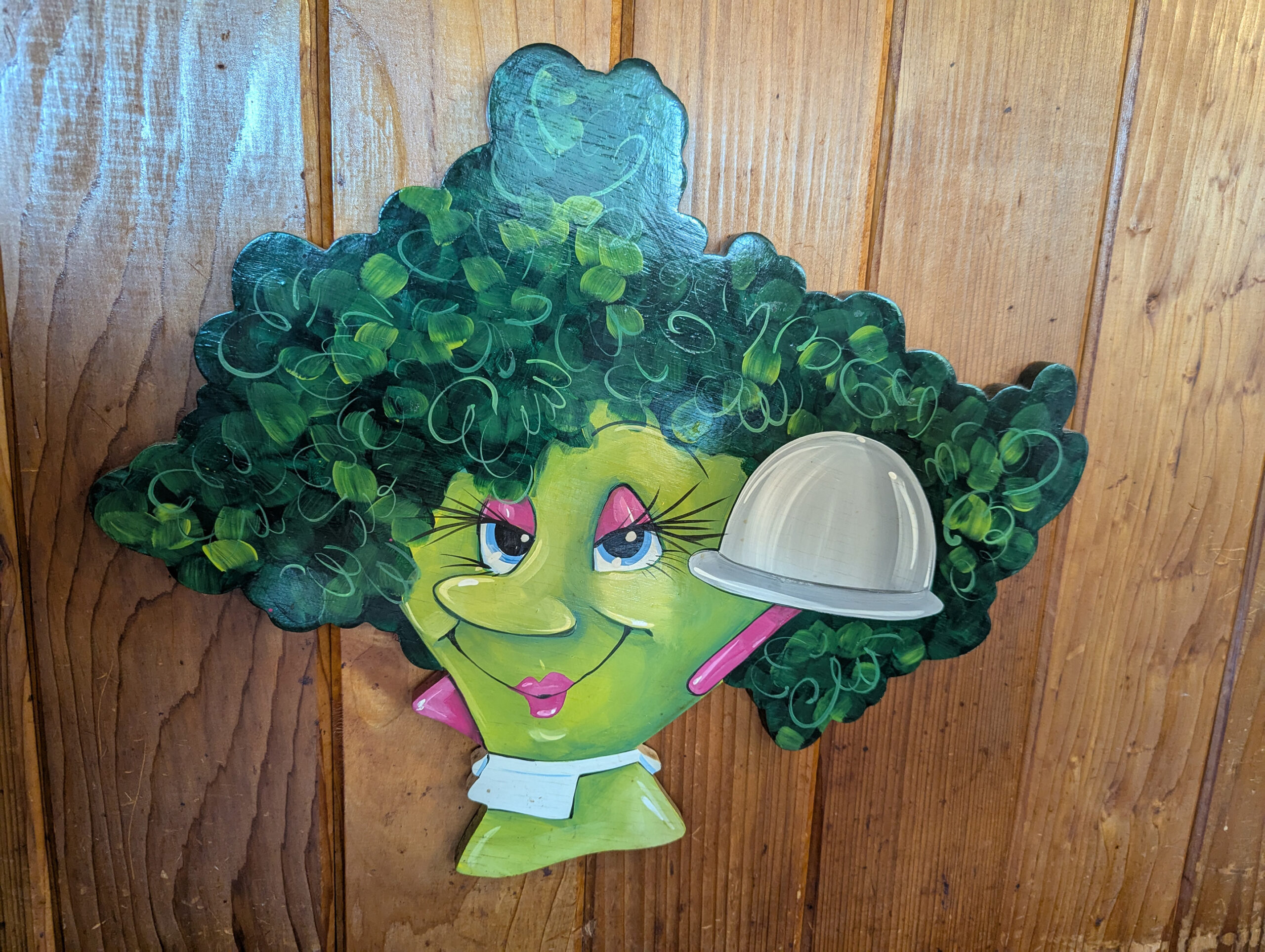 Painted piece of art on a wood-paneled wall. It is an caricature of a head of broccoli, anthropomorphized as a woman with long eyelashes, pink eye shadow, and pink lipstick and earrings. The leafy top of the broccoli is the hair.