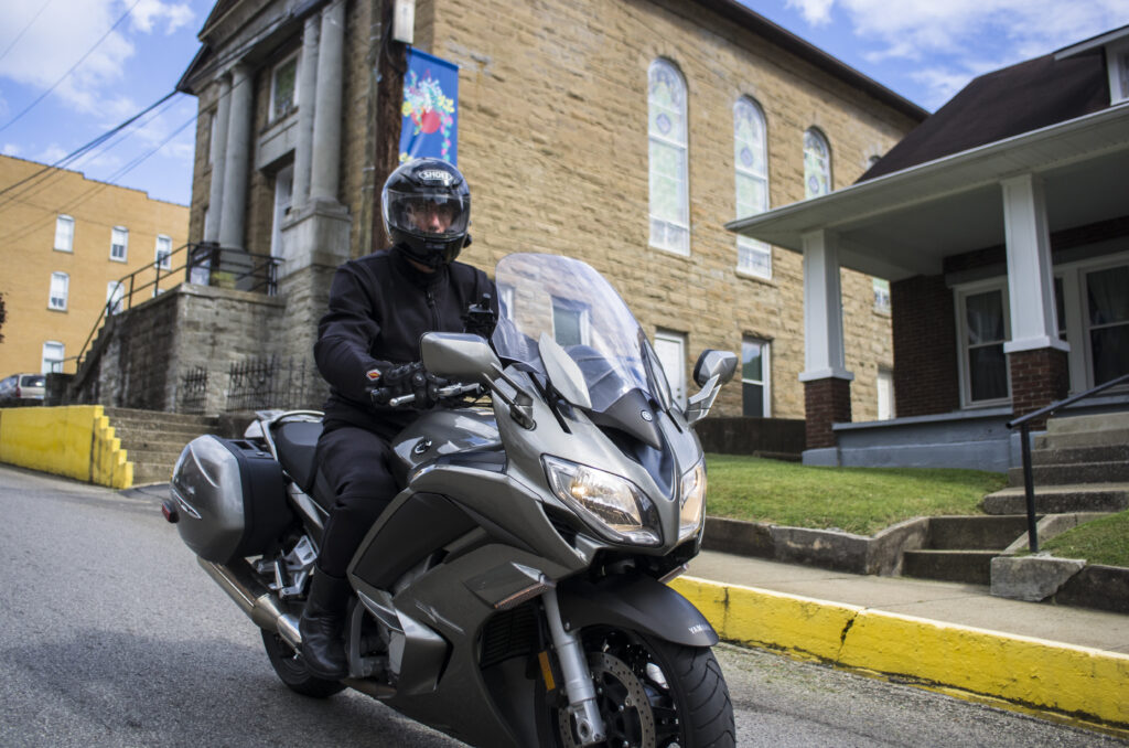 A man rides a touring motorcycle down a steep hill in a small town. There is a brick home and a stone church in the background.