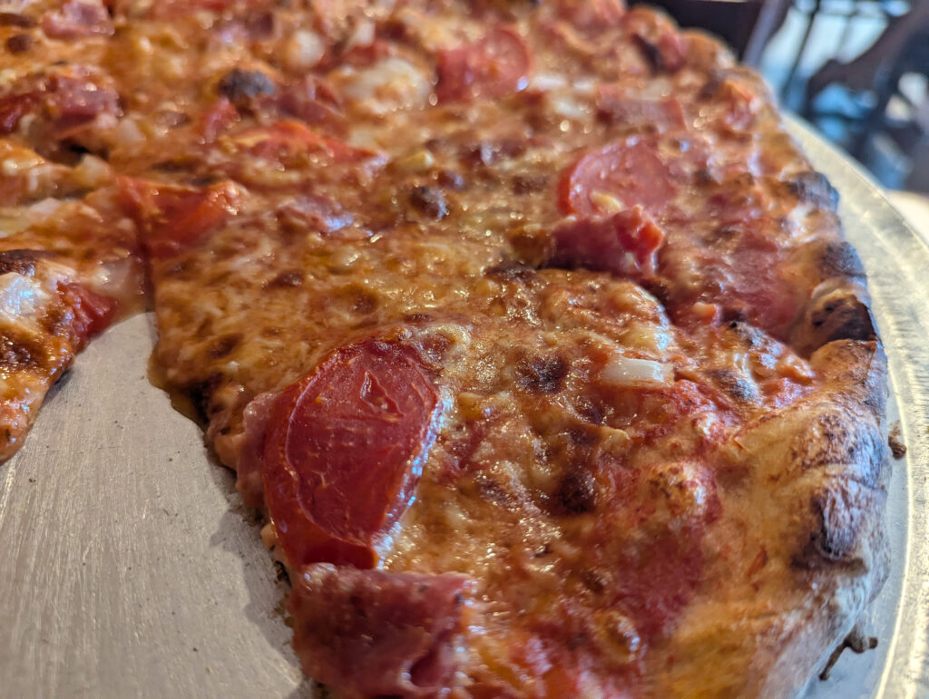 Photo of a pizza pie on a round aluminum serving tray with one slice of pizza removed. It's fresh from the oven, so the cheese is still melted. There are slices of tomato on top of the cheese along with smaller slices of salami