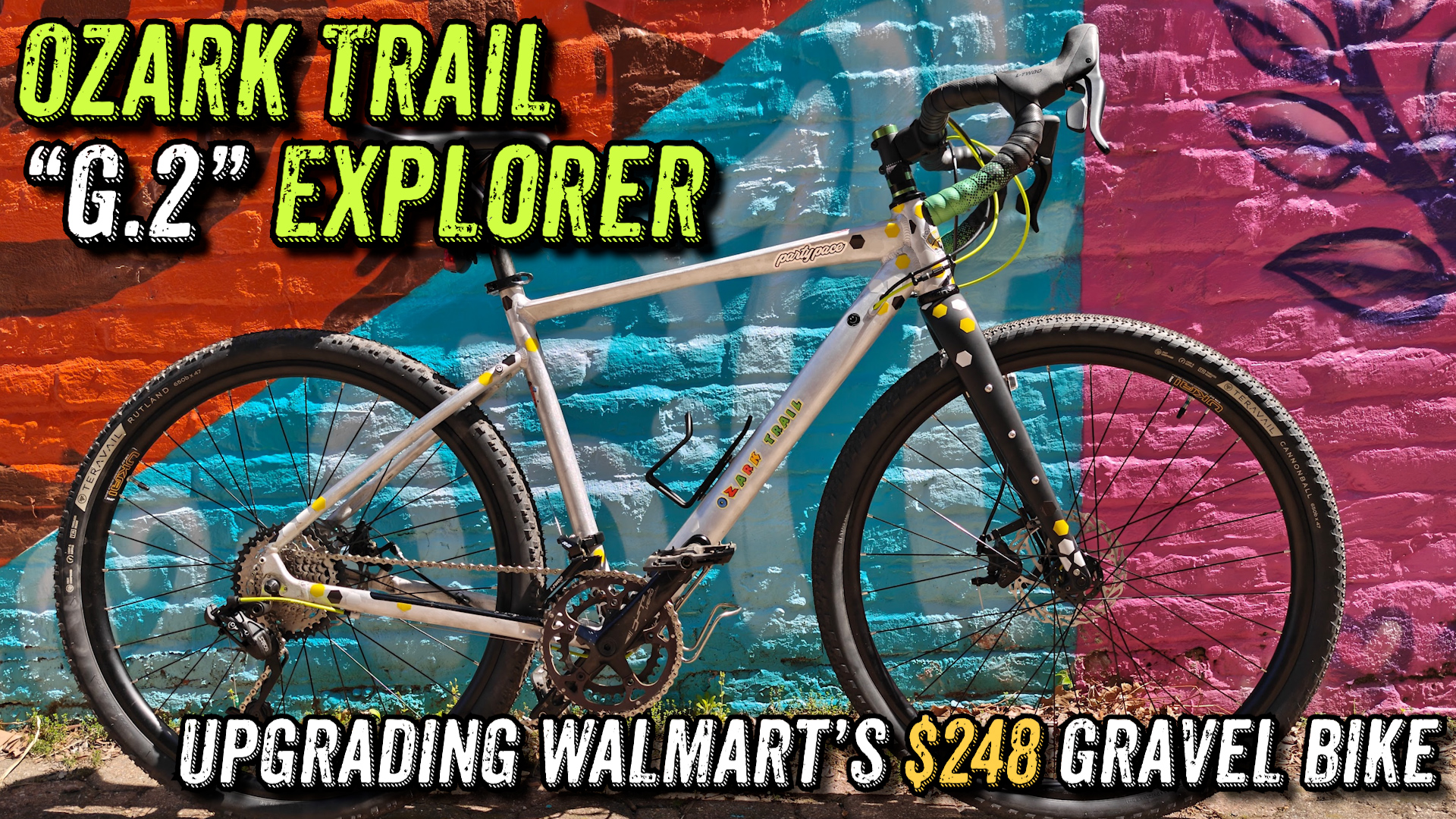Photo of a silver and black bicycle standing against a colorful brick wall mural. The mural has a large triangle of turquoise with a block of pink to the right and orange above. Within each of the color blocks are additional graphics in the hue of that block. Headline, "Ozark Trail 'G.2' Explorer" Subhead, "Upgrading Walmart's $248 Gravel Bike